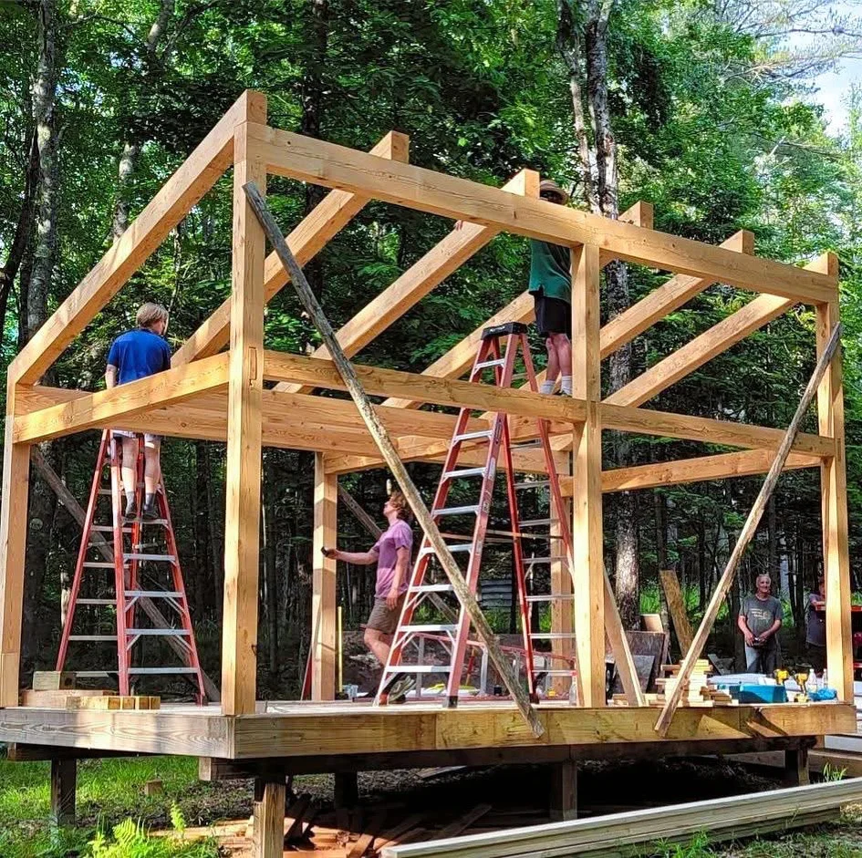 Timber frame pavilion under construction with exposed beams and diagonal bracing in wooded setting