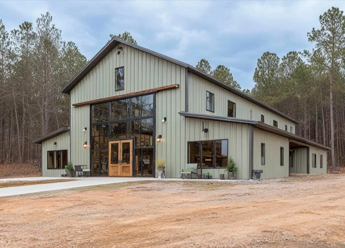 Modern barndominium with metal siding, large windows, and gable roof surrounded by pine trees