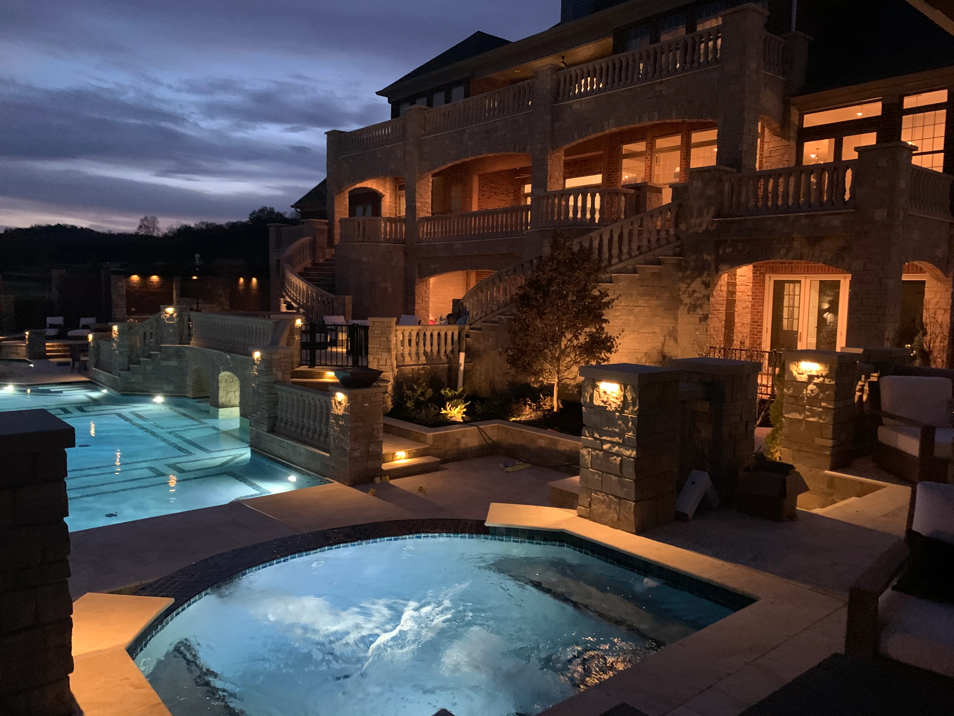 Illuminated luxury pool and spa at dusk with brick mansion and stone balustrades