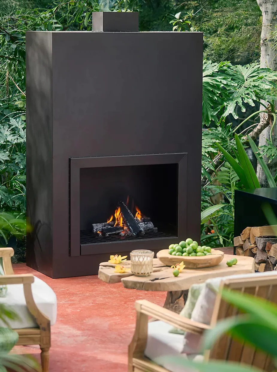 Modern black metal outdoor fireplace with active fire, surrounded by green plants and wooden furniture on terracotta patio